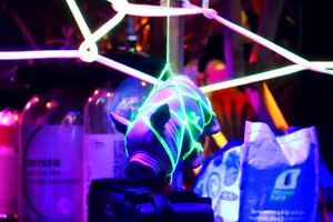 A scene at the bar.
A gummy pig is wrapped in green neon string shibari
and suspended in the center of the picture.
There are various colors and unfocused elements in the picture.