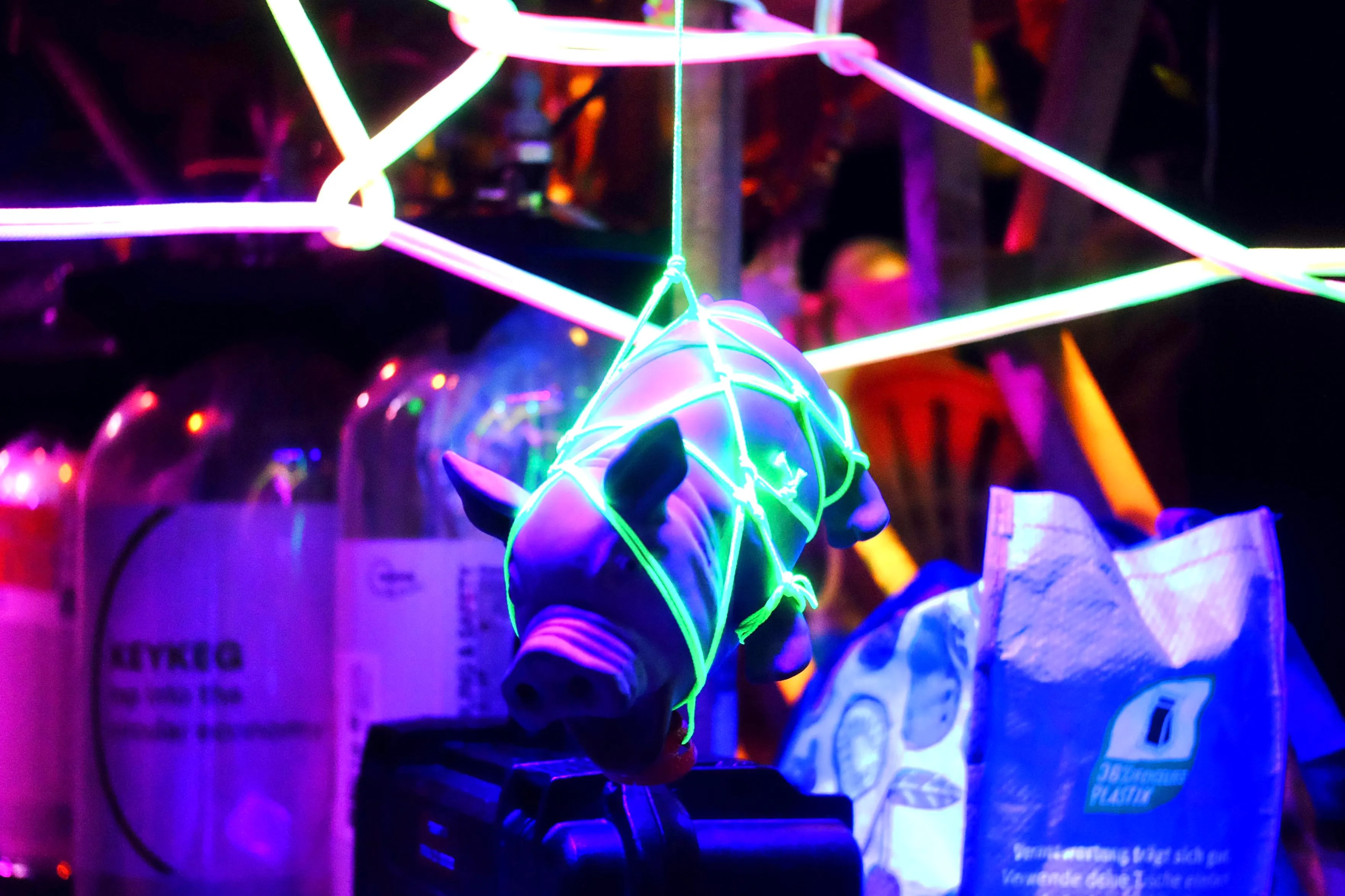 A scene at the bar.
A gummy pig is wrapped in green neon string shibari
and suspended in the center of the picture.
There are various colors and unfocused elements in the picture.