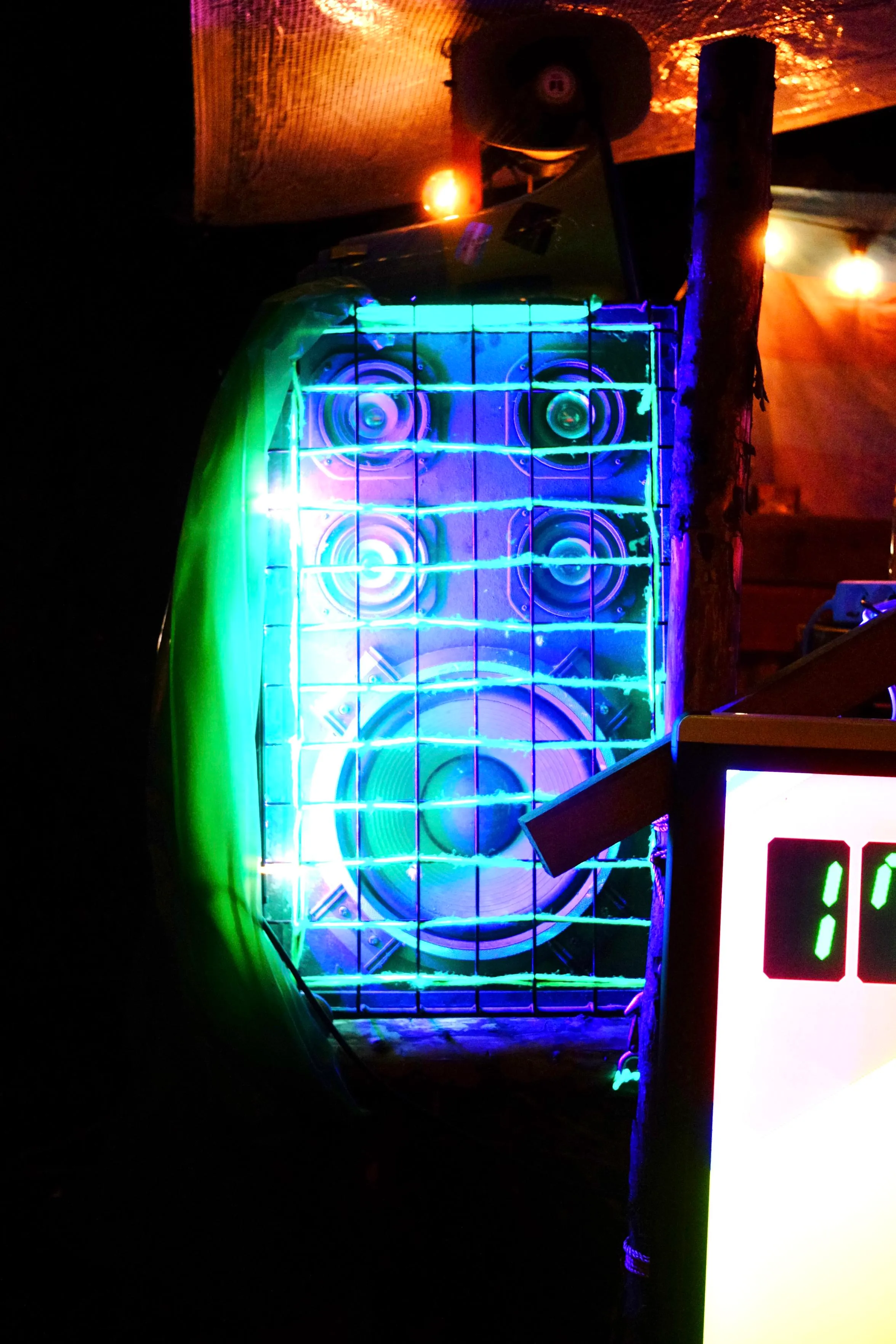 A speaker next to the DJ desk at the bar.
It's illuminated in green and violet and has a wire grid in front of it.
We can see a tarp above and in the background being lit
with shades of red, orange and yellow.
The DJ desk and pixelflut screen are extending to the right.