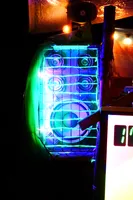 A speaker next to the DJ desk at the bar.
It's illuminated in green and violet and has a wire grid in front of it.
We can see a tarp above and in the background being lit
with shades of red, orange and yellow.
The DJ desk and pixelflut screen are extending to the right.