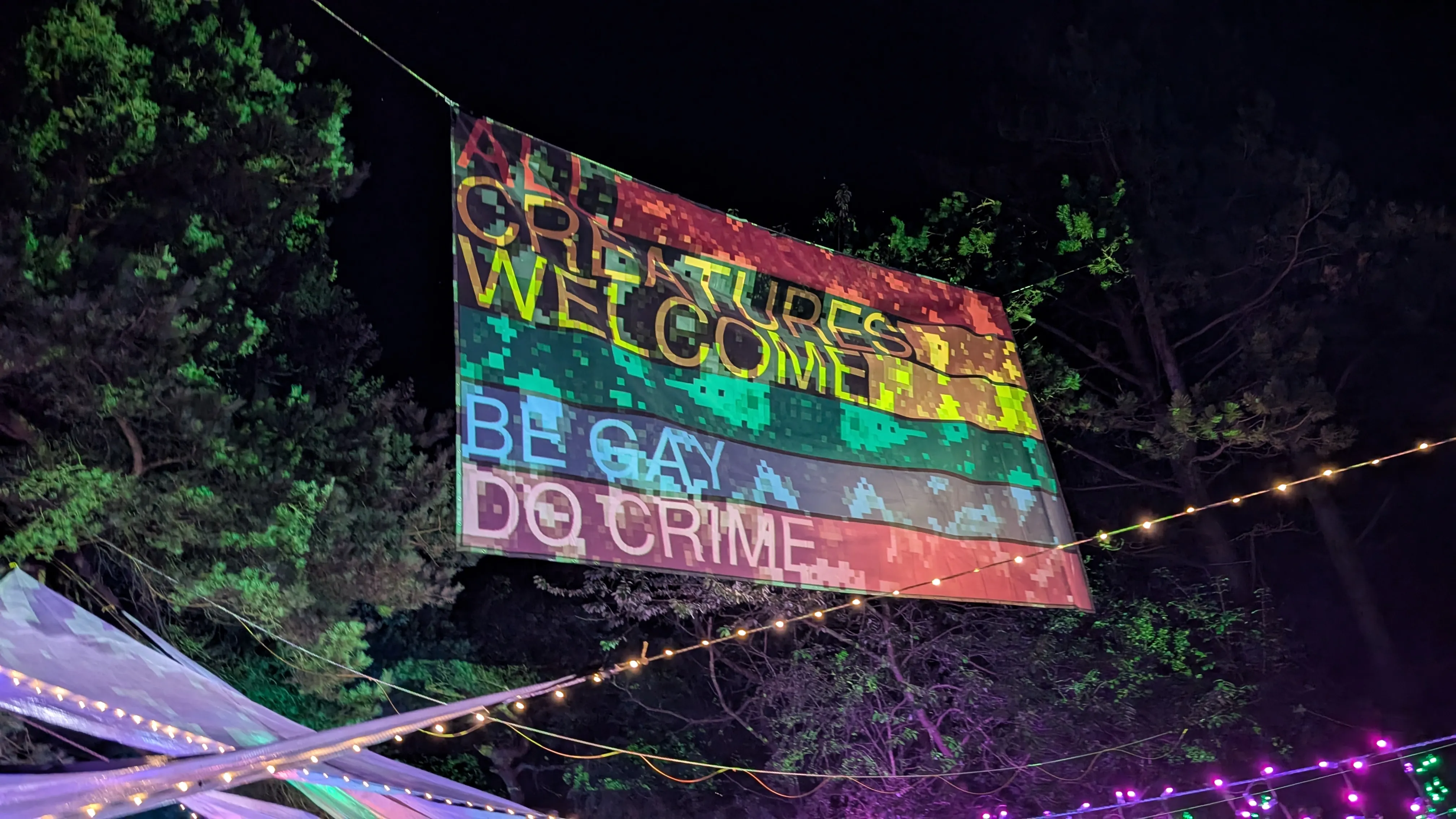A big, colorful banner hanging in the air over the bar area at night.
The banner is illuminated and colored in rainbow stripes.
We can see two sections of text on it
reading 'All creatures welcome' and 'be gay do crime'.
Underneath the banner we can see several string lights in different colors.