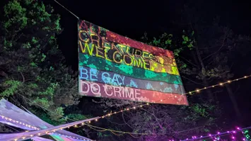 A big, colorful banner hanging in the air over the bar area at night.
The banner is illuminated and colored in rainbow stripes.
We can see two sections of text on it
reading 'All creatures welcome' and 'be gay do crime'.
Underneath the banner we can see several string lights in different colors.