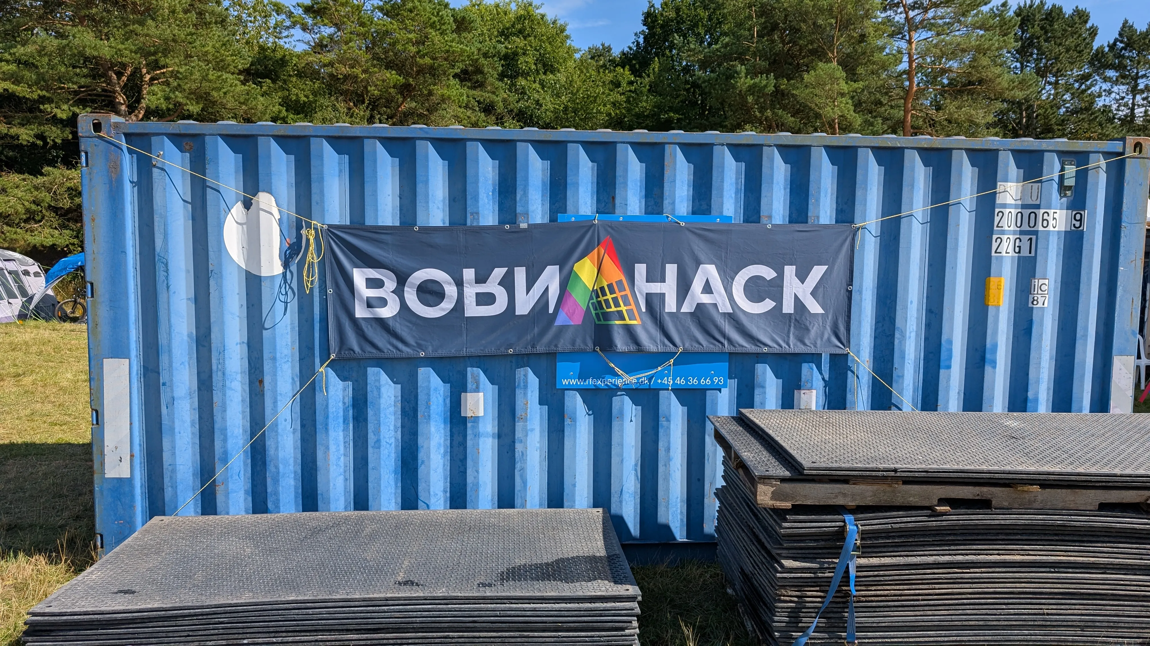 Photograph of the side of a blue shipping container.
It has a BornHack banner on it with the logo in pride colors.
We can see some greenery around the container
and there are some ground mats piled up in front of it.