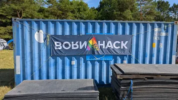 Photograph of the side of a blue shipping container.
It has a BornHack banner on it with the logo in pride colors.
We can see some greenery around the container
and there are some ground mats piled up in front of it.