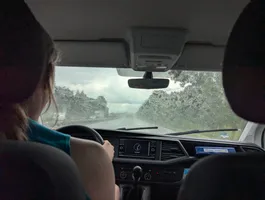 A picture taken from the back of a van towards the front window.
We can see Fiona's back as she's driving down a highway.
There's rain on the front window.
The sky is partially cloudy
and we can see some greenery on the sides of the road.