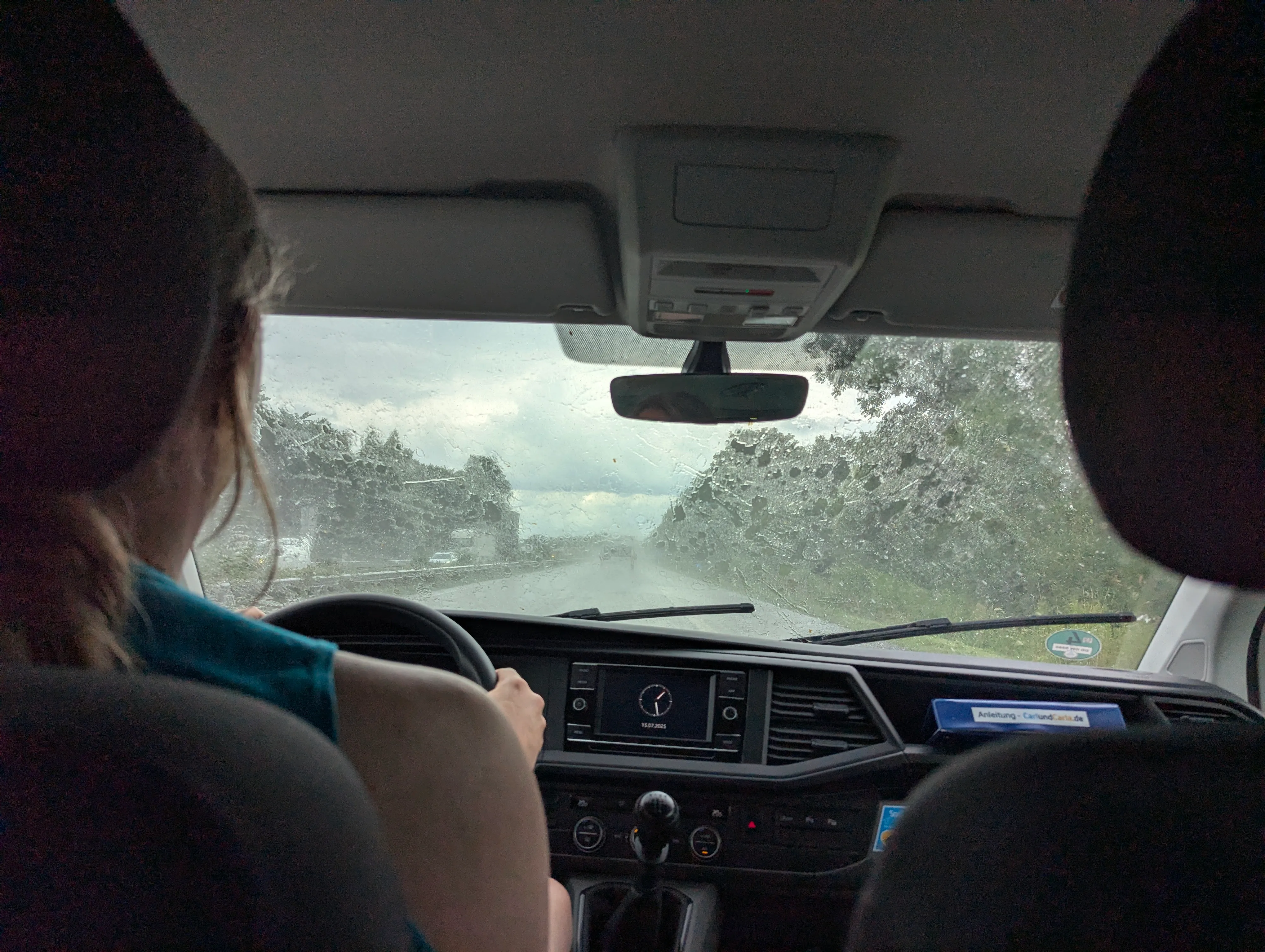 A picture taken from the back of a van towards the front window.
We can see Fiona's back as she's driving down a highway.
There's rain on the front window.
The sky is partially cloudy
and we can see some greenery on the sides of the road.