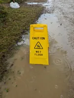 A warning sign reading 'caution - wet floor' is placed in a big puddle on the side of a dirt road.