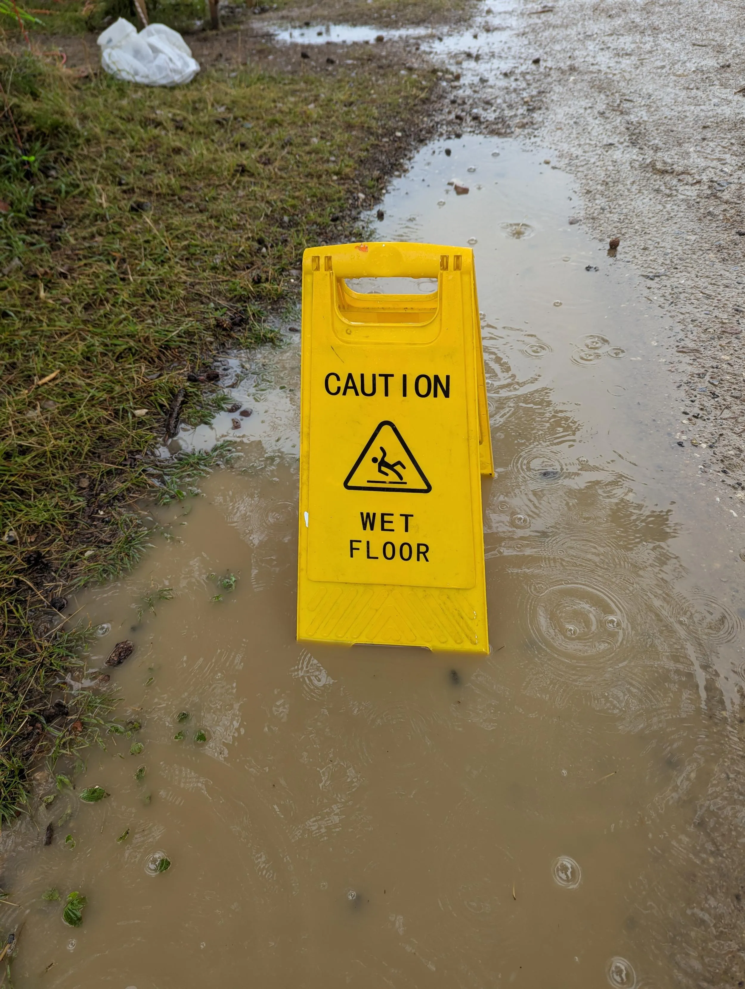 A warning sign reading 'caution - wet floor' is placed in a big puddle on the side of a dirt road.