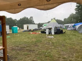 A view across parts of the general camping area towards the info desk.
We can see thick drops of rain pouring from a tarp on top of the picture.
There's a collection of empty trashcans lying on their sides under a star tent.