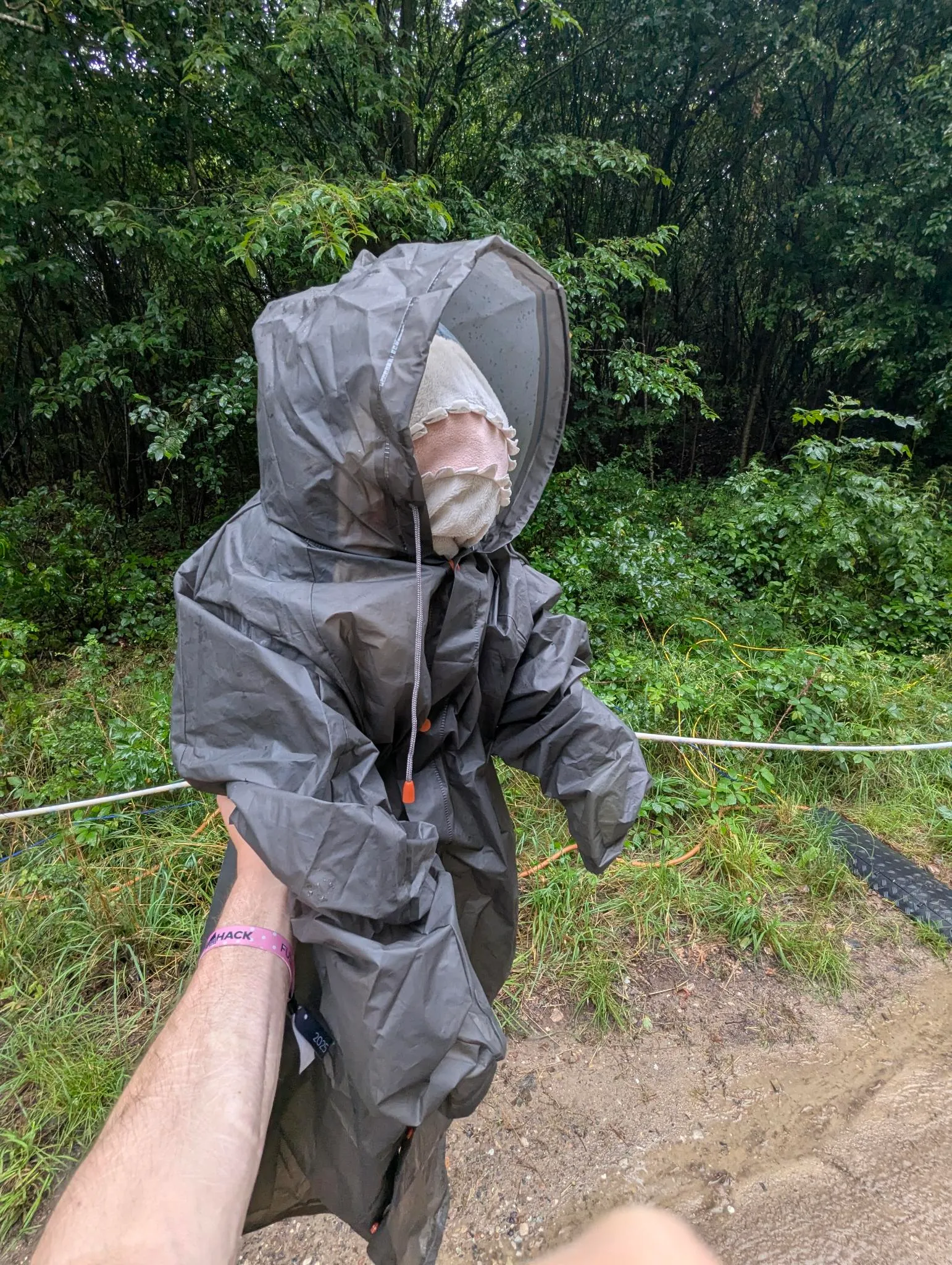 A blåhaj in a raincoat being held up at arms length in front of some greenery.