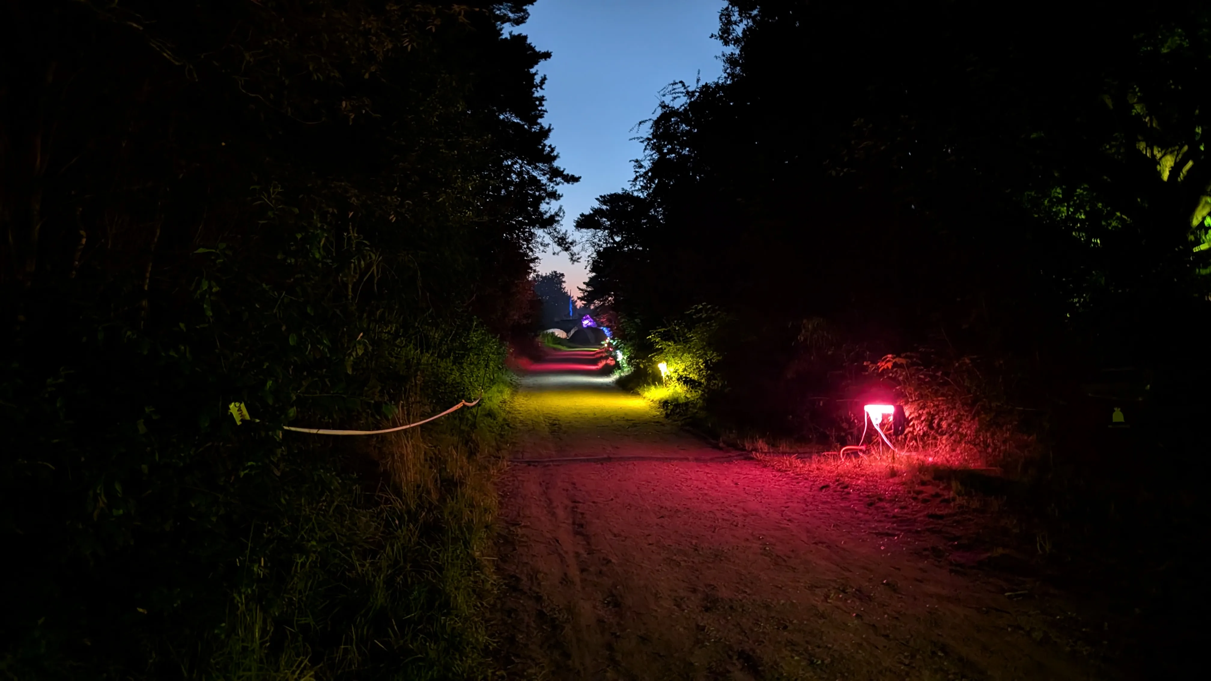 The main road around 4 in the morning.
We can see the empty road towards the general camping area
as it cuts a line through the trees.
Above the road we can see the sky fading to bright blue
with a tint of orange and yellow towards the horizon.
Colorful lights are placed at the road sides.