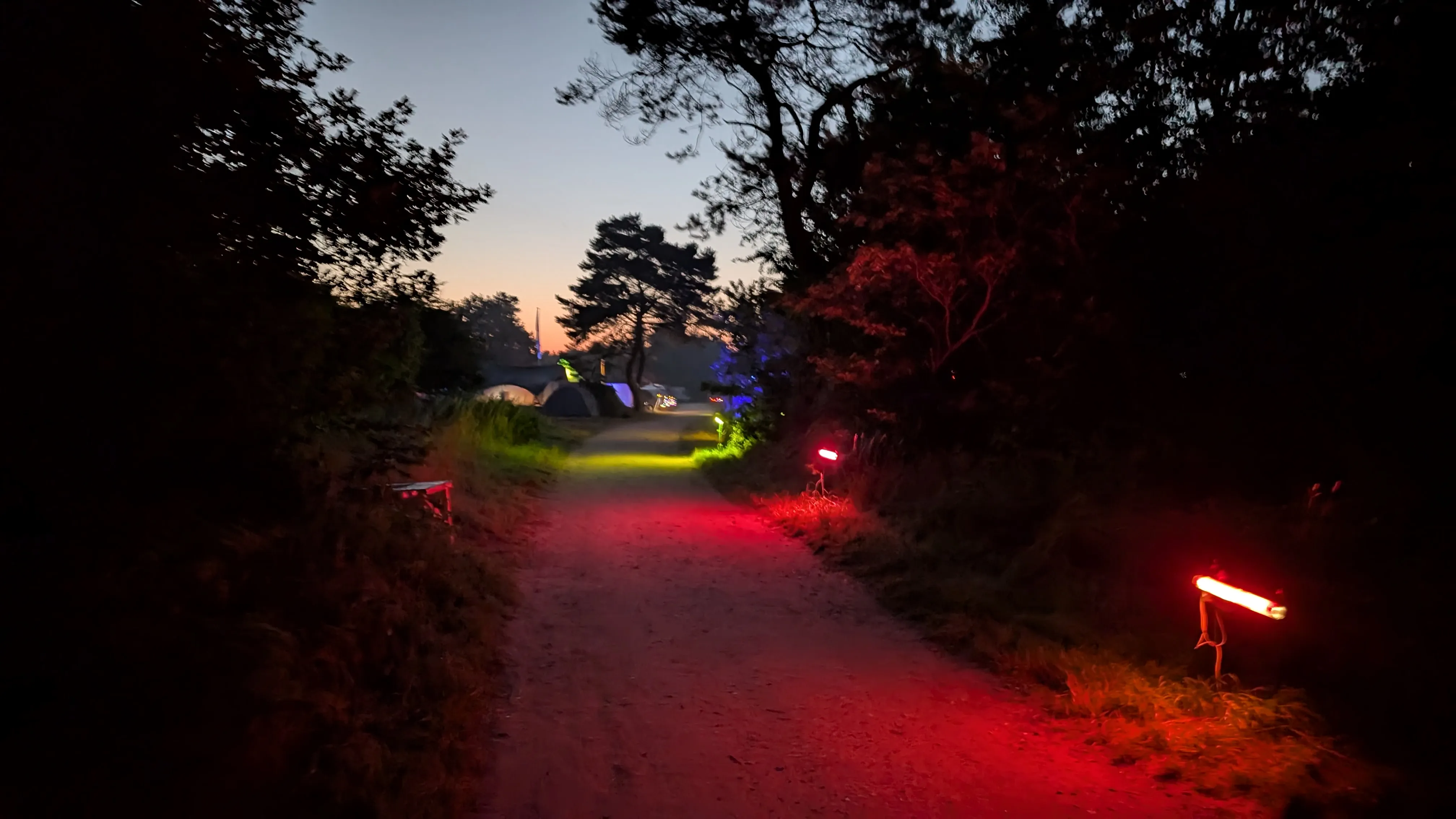 The main road around 4 in the morning.
The road opens up towards the general campsite here.
We can see a singular tree in the middle of the photo, surrounded by a number of tents.
The sun will clearly rise soon, as suggested by the sky.
There are still several colorful lights illuminating the path.