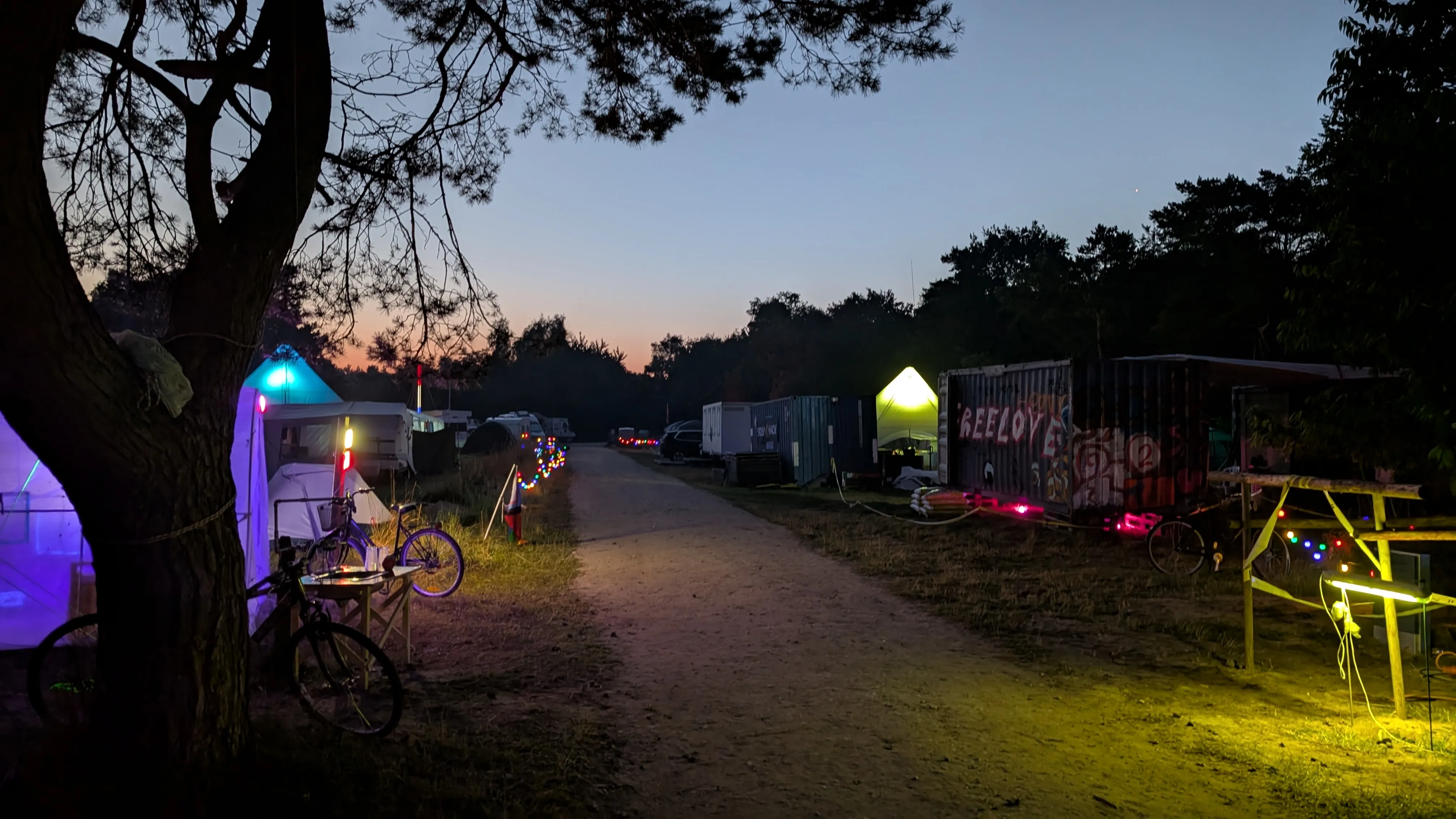 Early morning view down the main road through the general camping area.
We can see differently colored lights along the path as well as
a graffiti covered container with 'freelove' written on it.
The sun is starting to rise on the horizon.