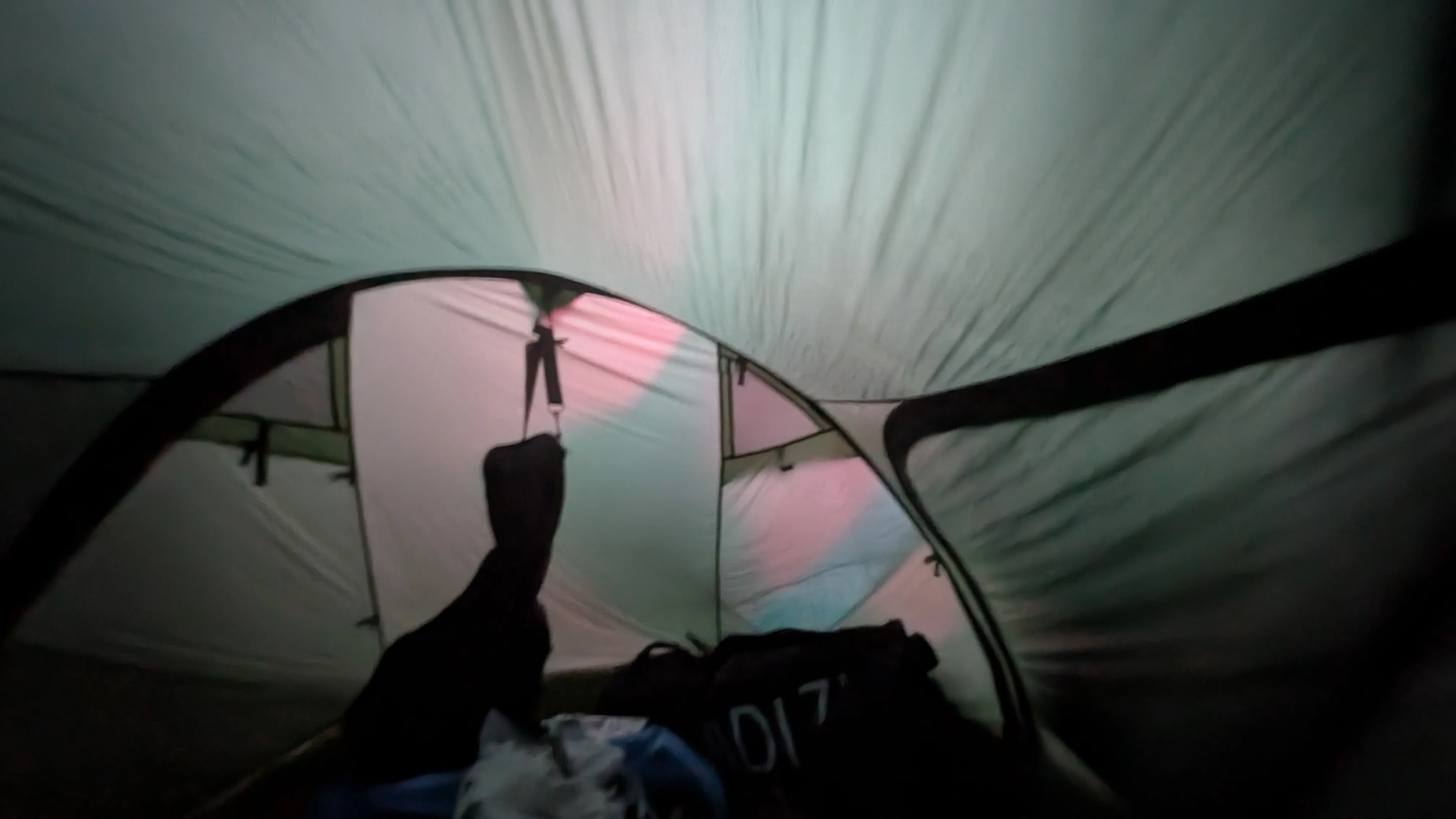 View from inside the tent.
We can see a trans flag casting is colors
onto the tent wall as the morning light filters through it.
The silhouette of a guitar bag visible against the tent wall.