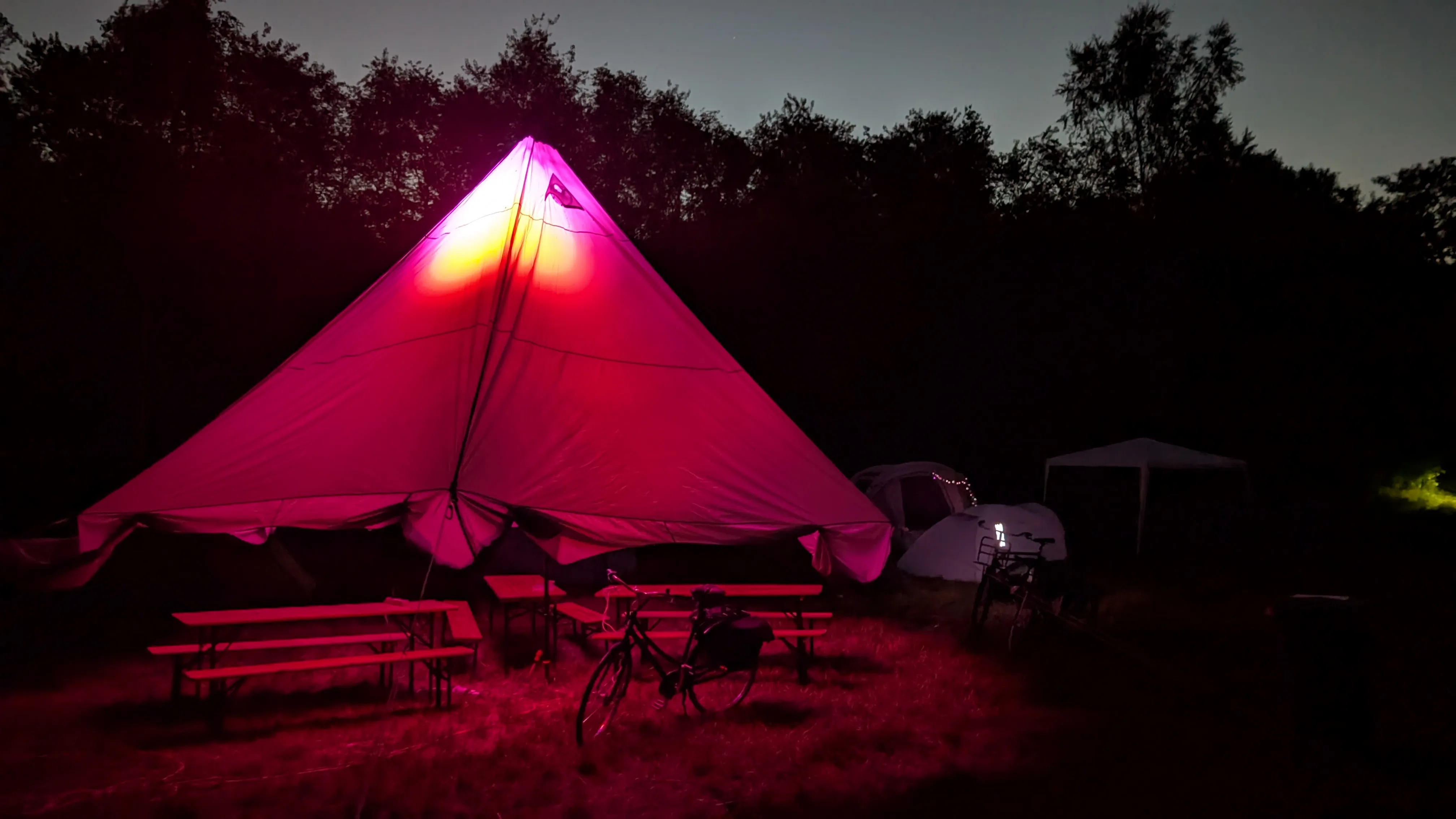 The community tent at cozy at night.
The tent is illuminated with red and pink light.
We can see some benches and tables under the tent.
We can see the sky being lighted in a soft grey against the dark treeline.