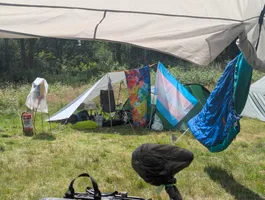 View from the community tent at the cozy camping area towards the tent were Fiona and her partner slept.
We can see that the distance between the power distribution point and the tent is now bridged by a tarp with a chair,
table and some stuff under it.
Attached to the tarp we can see a colorful towel and a trans flag.
In the background we can see some tall gras and a treeline.